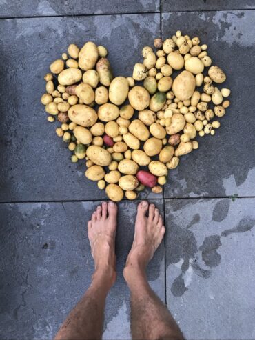 Verschiedene frisch geerntete Kartoffeln liegen herzförmig auf dem nassen Bürgersteig, und am unteren Rand des Bildes sind die nackten, schmutzigen Füße einer Person zu sehen.