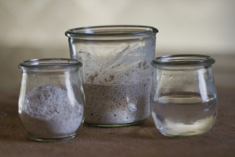 Drei kleine Gläser werden auf eine Fläche gestellt: Eines enthält Mehl, ein anderes einen aktiven Sauerteigstarter und das dritte ist mit Wasser gefüllt. Die Gläser sind durchsichtig und haben keine Etiketten.