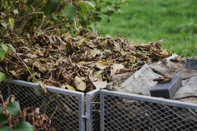 Eine mit trockenem Laub und Gartenabfällen gefüllte Komposttonne steht im Freien auf grünem Gras, teilweise abgedeckt mit einem Jutesack und einem schwarzen Ziegelstein. Um die Tonne herum sind Sträucher zu sehen.