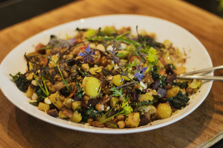 Ein farbenfroher Salat auf einem weißen Teller mit Kichererbsen, gelben Kirschtomaten, Grünzeug, Gemüsewürfeln, Samen und essbaren Blüten, der auf einer hölzernen Unterlage mit Servierutensilien angerichtet ist.