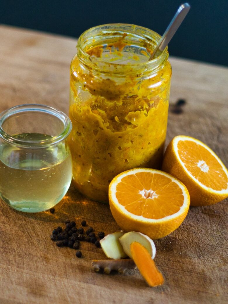 Ein Glas mit orange-gelber Paste, ein Löffel, ein kleines Glas mit Flüssigkeit, eine halbierte Orange, schwarze Pfefferkörner, Ingwer und Kurkumawurzelscheiben werden auf einer hölzernen Unterlage angeordnet.