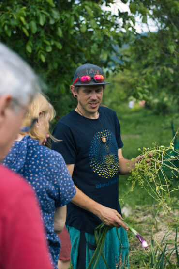 Ein Mann mit blauem T-Shirt, Mütze und rosafarbener Sonnenbrille auf dem Kopf steht im Freien, hält frisch gepflückte grüne Zwiebeln in der Hand und spricht mit zwei Personen im Vordergrund. Im Hintergrund sind Bäume und grünes Gras zu sehen.