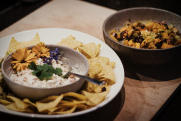 Ein Teller mit Tortilla-Chips und einer Schale mit cremigem Dip, garniert mit essbaren Blüten und Koriander, daneben eine Schale mit gewürfeltem, geröstetem Gemüse auf einer Holzplatte.