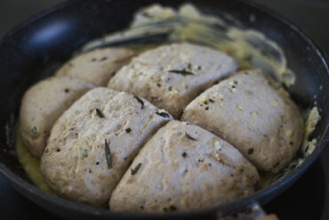 Sechs Stück ungebackener Kräuterbrotteig liegen in einer runden Pfanne, bestrichen mit Butter und Kräutern, bereit zum Backen.