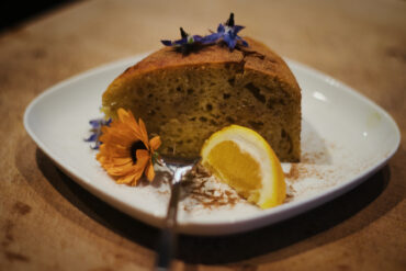 Ein Stück Kuchen auf einem weißen Teller, garniert mit einer Zitronenspalte, essbaren Blüten und einer Prise Zimt, wobei eine Gabel auf dem Teller liegt.