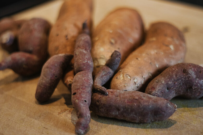 Mehrere Süßkartoffeln unterschiedlicher Größe und Form mit rötlich-brauner Schale liegen auf einer Holzfläche. Der Fokus liegt auf den Süßkartoffeln im Vordergrund.