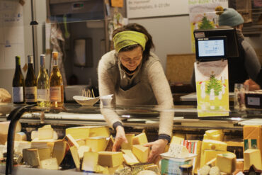 Eine lächelnde Frau mit grünem Stirnband arrangiert Käseblöcke an einem Marktstand, auf dessen Tresen Weinflaschen und eine Digitalwaage zu sehen sind.