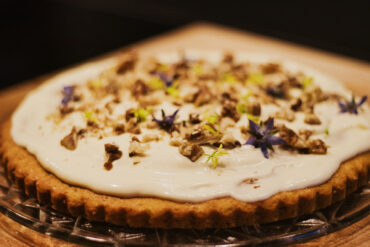Eine runde Torte mit goldener Kruste, die mit cremig-weißem Zuckerguss, gehackten Nüssen und kleinen lilafarbenen essbaren Blüten belegt ist und auf einer Glasplatte vor einem dunklen Hintergrund präsentiert wird.