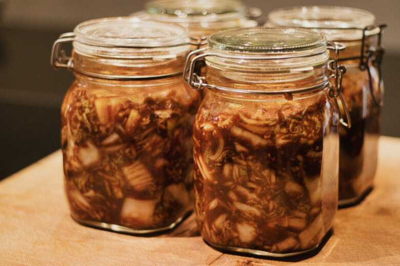 Vier Gläser, gefüllt mit hausgemachtem Kimchi, mit gehacktem Gemüse und einer rötlich-braunen, würzigen Lake, sind auf einer Holzfläche angeordnet. Die Gläser sind mit Metallverschlüssen und klaren Deckeln verschlossen.