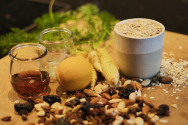 A wooden board with a cup of oats, a bunch of yellow carrots, a lemon, a jar of honey, an empty glass jar, and a scattered mix of nuts and dried fruits. Green carrot tops are in the background.