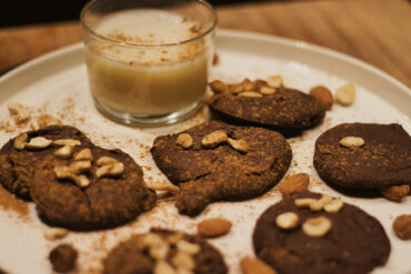A white plate with chocolate cookies topped with cashews and almonds, sprinkled with cinnamon, next to a small glass of creamy drink.