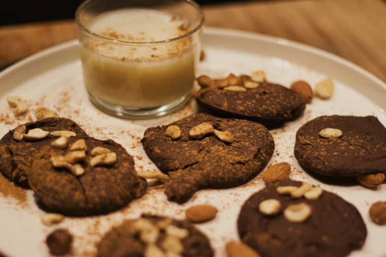 A white plate with chocolate cookies topped with cashews and almonds, sprinkled with cinnamon, next to a small glass of creamy drink.