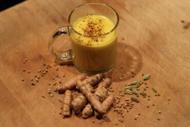 A glass mug filled with yellow turmeric milk, sprinkled with spice on top, sits on a wooden surface next to fresh turmeric roots, coriander seeds, and cardamom pods.