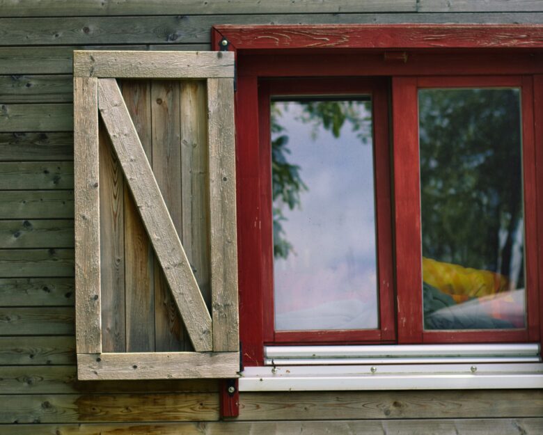 A close-up of a wooden building exterior shows a window with a bright red frame and a partially open, rustic wooden shutter. The window reflects trees and sky. The wall is made of horizontal wooden planks.