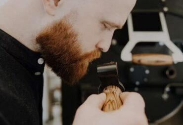 A bearded man closely smelling coffee beans held in a wooden scoop, with coffee roasting equipment visible in the background.