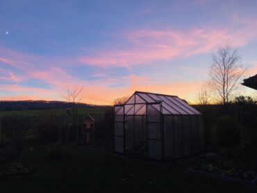 A glass greenhouse stands in a garden at sunset, with a pink and purple sky, distant hills, bare trees, and a crescent moon visible. The scene has a peaceful, rural atmosphere.