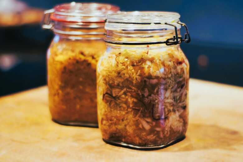 Two glass jars filled with shredded, fermented vegetables, likely sauerkraut or kimchi, are sitting on a wooden surface with a blurred background.