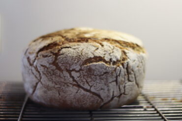 A round loaf of rustic, crusty bread with a cracked, flour-dusted surface sits on a metal cooling rack against a plain background.