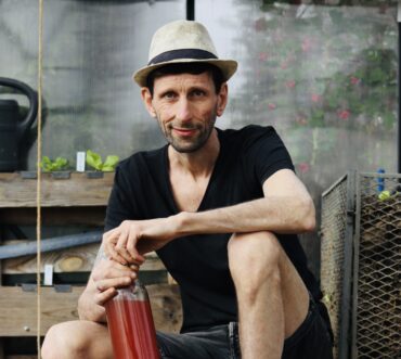 A man in a straw hat and black shirt sits outdoors, holding a bottle of reddish liquid. He is sitting on one knee in front of a garden setup with plants and wooden structures.