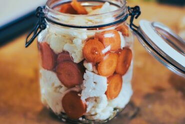 A glass jar filled with sliced carrots and cauliflower, with the lid open, sitting on a wooden surface.