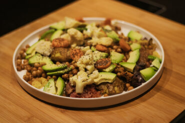 A white plate on a wooden surface holds a colorful dish with chickpeas, sliced zucchini, cauliflower, carrots, and other assorted vegetables, topped with seeds or seasoning.