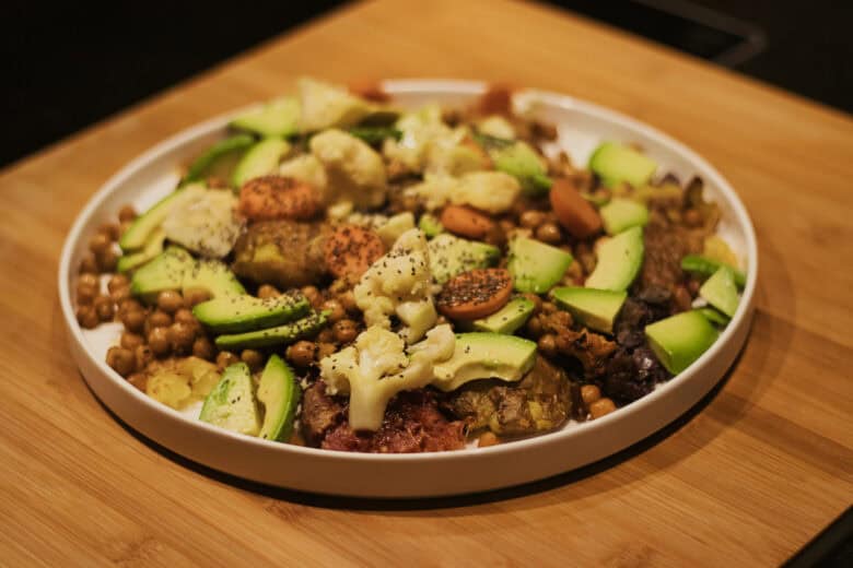 A white plate on a wooden surface holds a colorful dish with chickpeas, sliced zucchini, cauliflower, carrots, and other assorted vegetables, topped with seeds or seasoning.