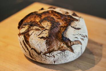 A round loaf of rustic sourdough bread with a crisp, golden-brown crust and flour-dusted surface sits on a wooden cutting board.