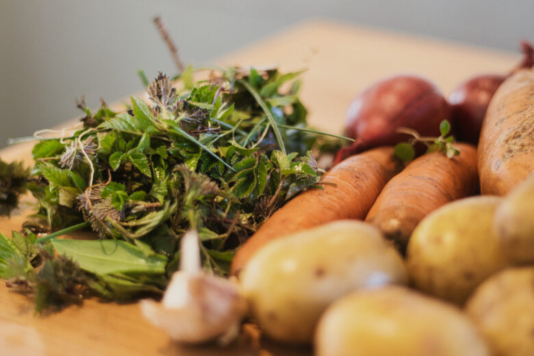 A close-up of fresh vegetables on a wooden surface, including potatoes, carrots, red onions, garlic, and a pile of leafy greens. The focus is on the foreground, with a soft background blur.