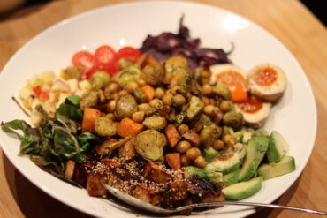 A colorful bowl filled with assorted roasted vegetables, avocado slices, leafy greens, cherry tomatoes, purple cabbage, chickpeas, tofu, and two halved eggs with sesame seeds, on a light wooden surface.