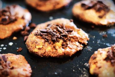 A close-up of freshly baked chocolate chip cookies on a dark baking tray, topped with chunks of melted chocolate. The cookies look soft and chewy with visible pieces of chocolate and nuts.