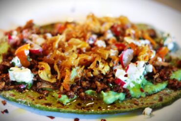 A close-up of a vibrant dish featuring a green flatbread topped with mashed avocado, crumbled cheese, chopped radishes, shredded vegetables, and crispy bits, all drizzled with olive oil on a white plate.