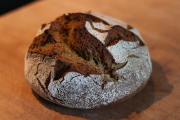 A round loaf of rustic, crusty bread with a golden-brown, deeply scored top sits on a wooden surface; the bread is dusted with flour and has a homemade, artisan appearance.