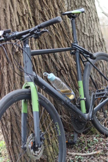 A black and green bicycle is leaning against a large tree trunk. A transparent water bottle with a blue cap is attached to the bike frame. The ground is covered with grass and small plants.