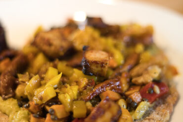 Close-up of a colorful dish featuring diced vegetables, grilled tofu or seitan, and possibly lentils or seeds, arranged on a flatbread or toast, with a shallow depth of field and a soft, blurry background.