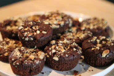 A plate of chocolate muffins topped with chopped nuts is arranged on a light-colored surface, with the focus on the texture and nutty topping of the muffins.
