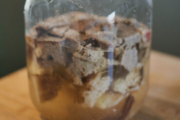 A glass jar filled with chunks of bread soaking in murky liquid, sitting on a wooden surface.