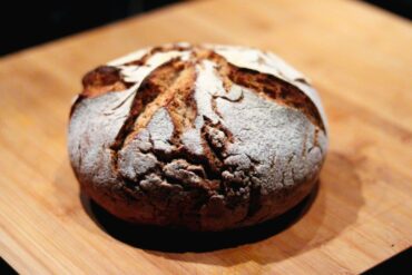 A round loaf of rustic sourdough bread with a golden-brown, crusty exterior and decorative scoring sits on a wooden surface.