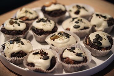 A plate of chocolate cupcakes with white frosting, topped with pumpkin seeds and nuts, arranged closely together in paper liners on a white tray.