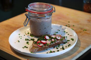 A slice of bread topped with pâté, sliced radishes, and chopped chives sits on a white plate, next to a glass jar filled with pâté, all placed on a wooden surface.