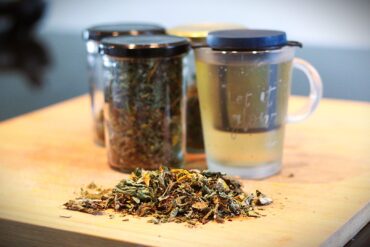 A pile of loose dried herbs sits on a wooden surface in front of three glass jars of dried herbs and a clear mug filled with herbal tea. The mug has black text on it. The background is blurred.