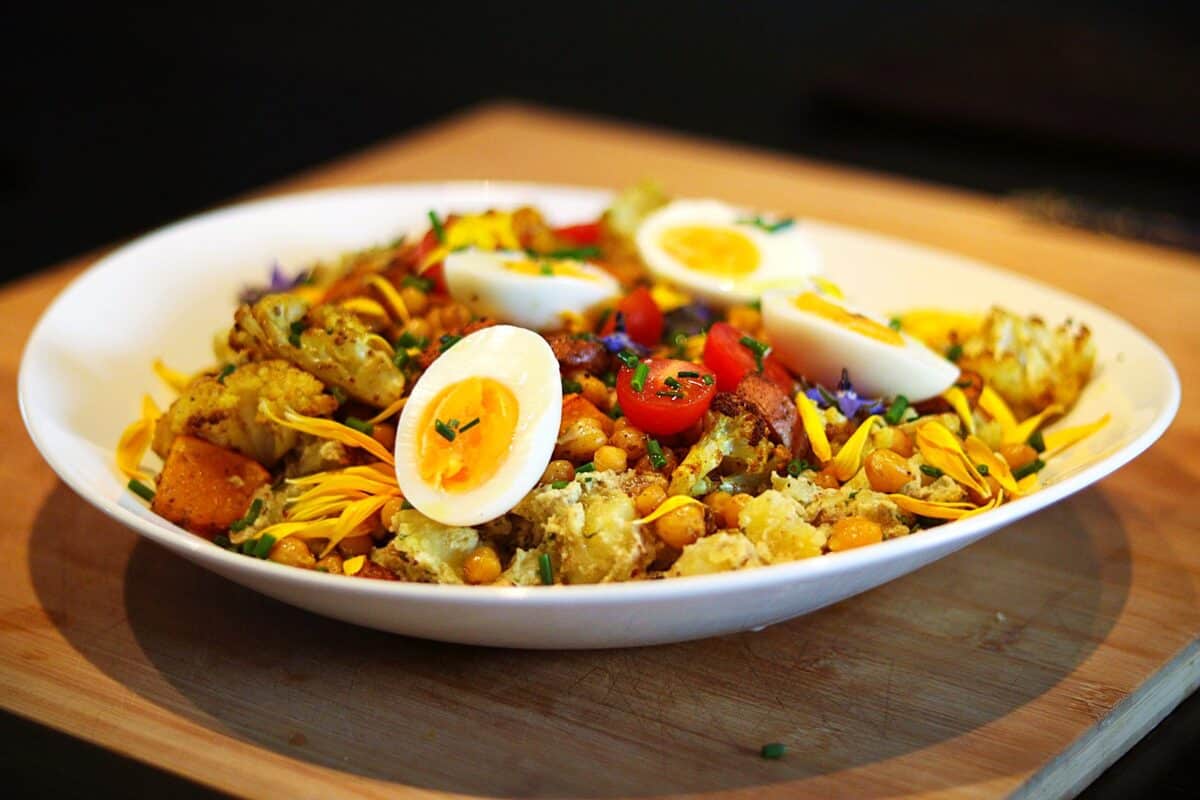 A white plate with roasted vegetables, chickpeas, halved boiled eggs, cherry tomatoes, and shredded yellow cheese, garnished with herbs, on a wooden surface.