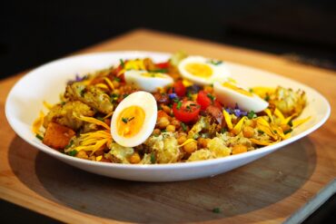 A white plate with roasted vegetables, chickpeas, halved boiled eggs, cherry tomatoes, and shredded yellow cheese, garnished with herbs, on a wooden surface.