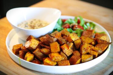 A white plate with roasted seasoned sweet potato cubes, a small green salad with cherry tomatoes, and a bowl of creamy dipping sauce on a wooden surface.