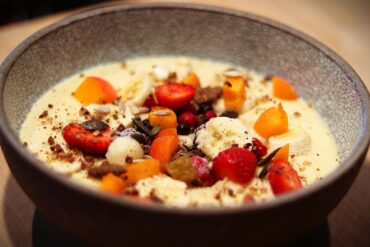 A bowl of creamy yogurt topped with fresh strawberries, banana slices, berries, chopped apricots, seeds, and crumbled nuts, served in a textured gray ceramic bowl.