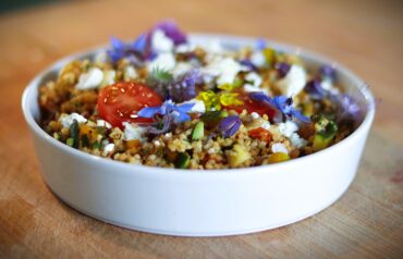 A white bowl filled with couscous salad, topped with cherry tomato slices, crumbled feta, chopped vegetables, and edible purple flowers, set on a wooden surface.
