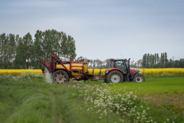 A red tractor with a yellow sprayer attached is driving through a green field, spraying crops. Yellow flowers and tall trees are visible in the background under a cloudy sky.