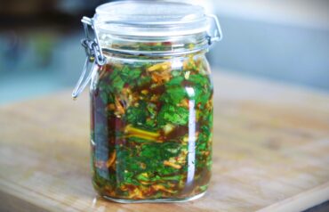 A glass jar with a clamp lid is filled with chopped fresh green herbs and vegetables in a dark liquid marinade, sitting on a wooden surface.