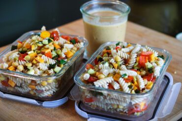 Two glass containers filled with colorful pasta salad, featuring rotini, chopped vegetables, and herbs, sit on a wooden surface. Behind them is a jar of creamy dressing.