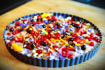A colorful tart in a fluted pan, topped with assorted berries, edible flowers, nuts, and seeds, sitting on a wooden surface.