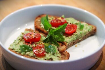 Two slices of whole grain toast topped with green spread, cherry tomato slices, fresh basil leaves, and herbs, served on a white plate.